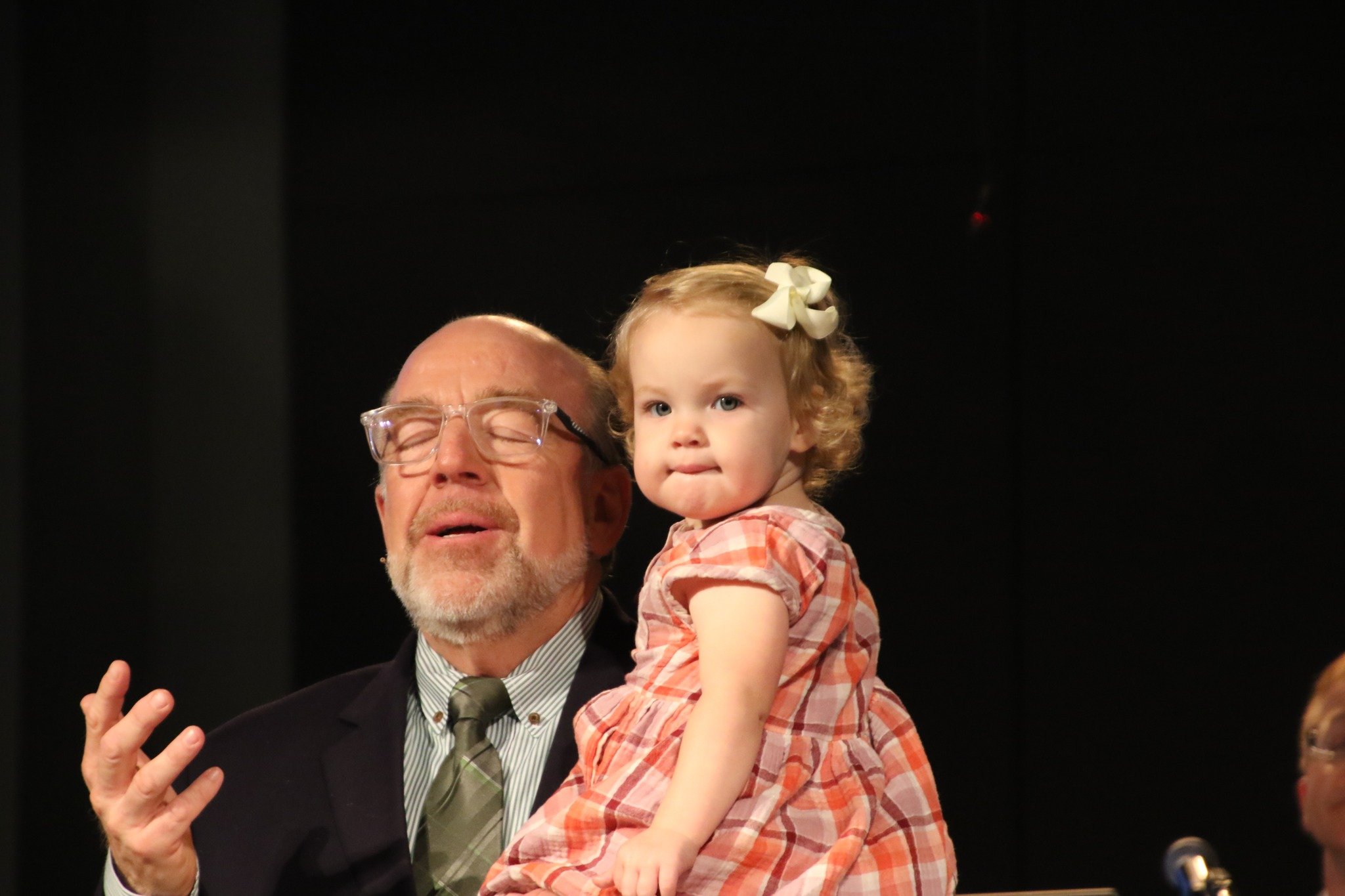 An elderly man with glasses and a gray beard, dressed in a suit and tie, holding a young girl with curly hair and a white bow, wearing a plaid dress, against a dark background.