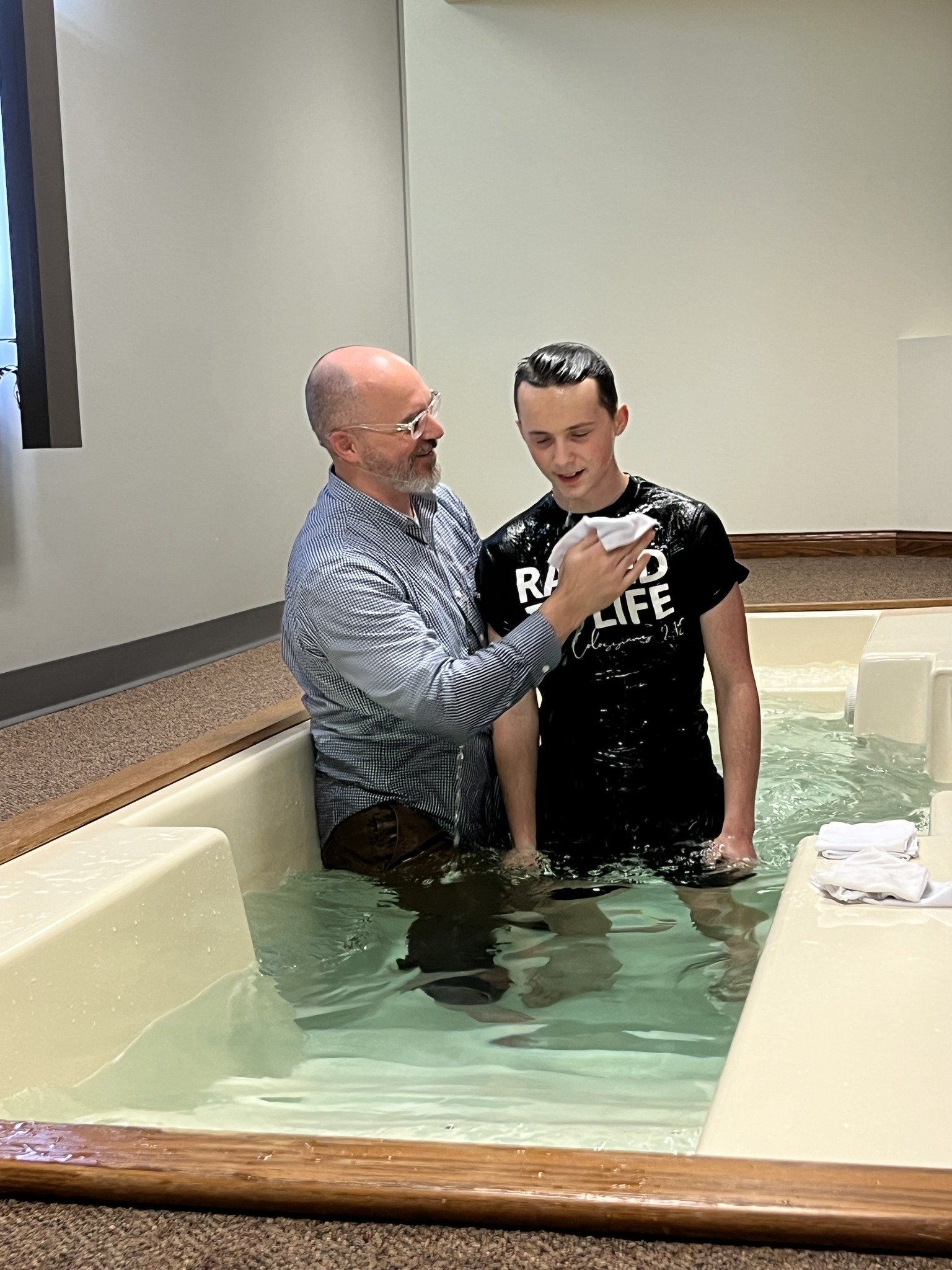 A man baptizing a young man in a baptismal pool, with water and white towels nearby.