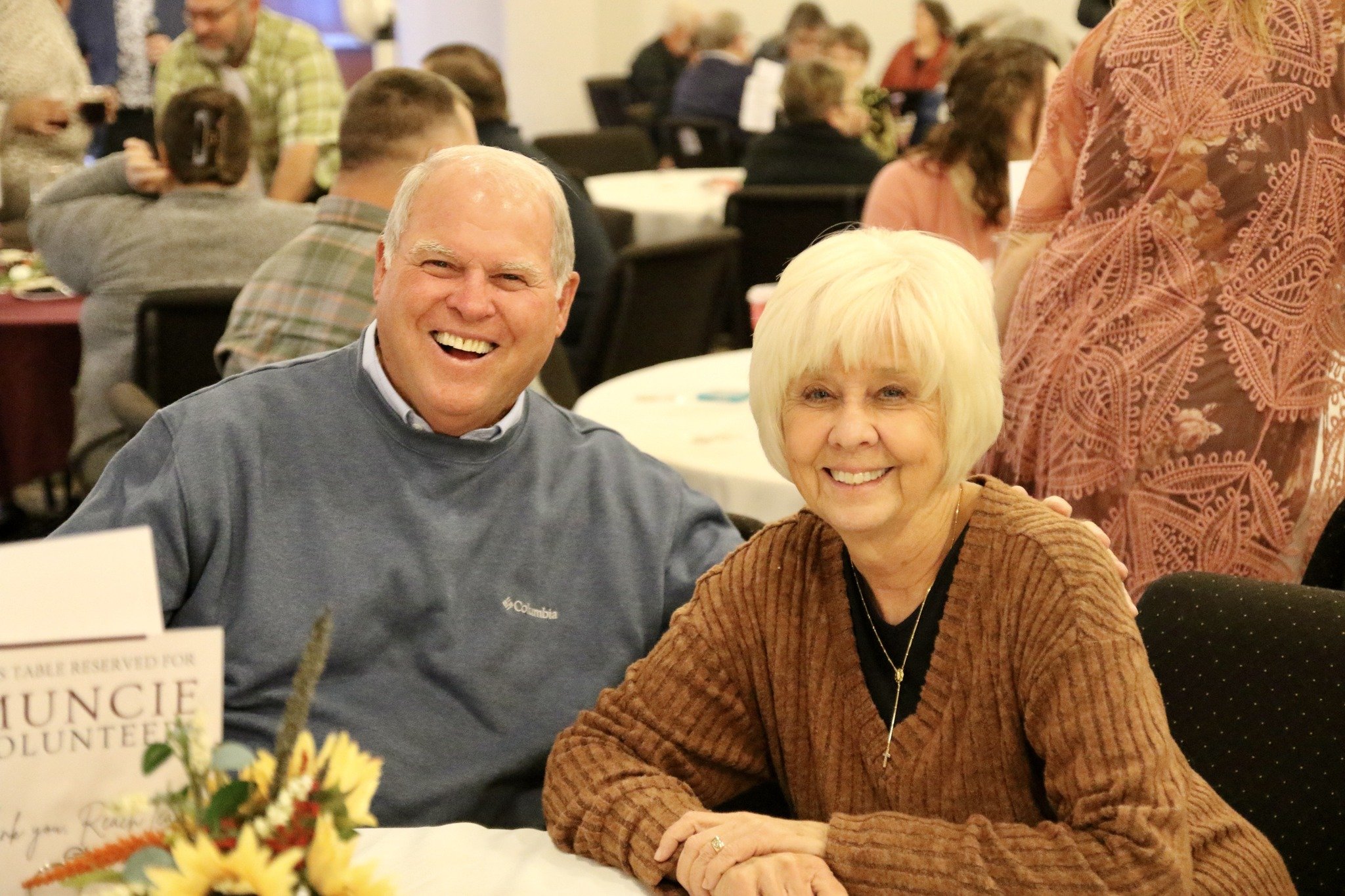 A senior man and woman smiling at a gathering or event, sitting at a table with a flower arrangement and a sign that says "Reserved for Muncie Volunteers."